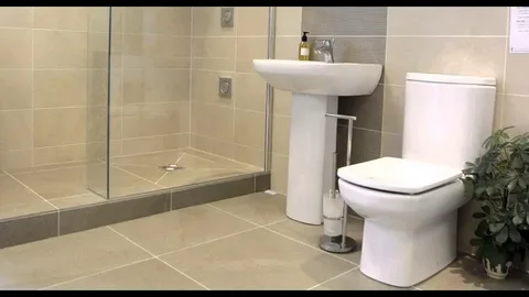 Best tile size for small bathroom displayed in a modern bathroom featuring a white pedestal sink, a white toilet, a glass-enclosed walk-in shower, beige tiled walls, and a small potted plant.