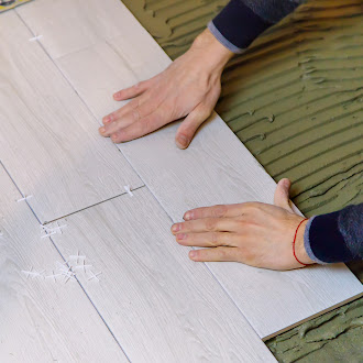 A person laying down light-colored wooden flooring tile installation on a textured green surface, carefully aligning the edges.