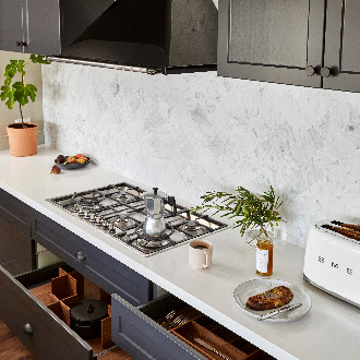 Modern kitchen featuring dark grey cabinets, sleek white countertop material, a gas stove, Smeg toaster, potted plant, and organized storage drawers for a functional and stylish design.