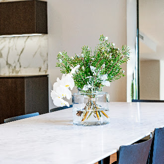Clear glass vase with white orchids and green foliage displayed on a white marble table, highlighting elegant countertop material choices in a modern kitchen with dark cabinets and marble walls.
