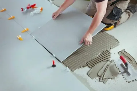 A person installs a large grey floor tile onto a bed of combed adhesive, using plastic spacers to ensure even spacing during the tiling process as part of professional ceramic tile maintenance and flooring installation.