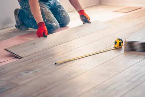 A worker in blue jeans and red gloves performs flooring installation services, laying light wood laminate flooring with a yellow tape measure resting on the planks in the foreground.