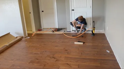 A person wearing knee pads performs flooring installation services, kneeling on a newly installed dark wood floor and using tools to finish the work near a white door in a bright room. 