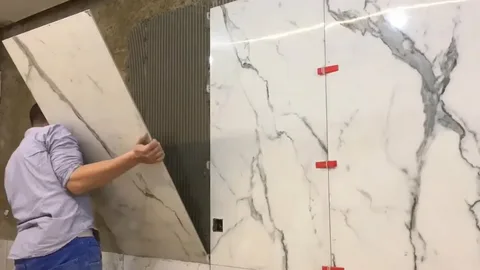 A person in a light shirt installs large white marble-patterned tiles onto a wall covered in adhesive, using red leveling clips between the panels during a professional ceramic tile maintenance and tile installation project.